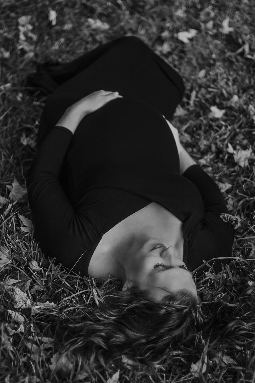 Black and white image of a pregnant woman in black dress lying on grass with fallen leaves.
