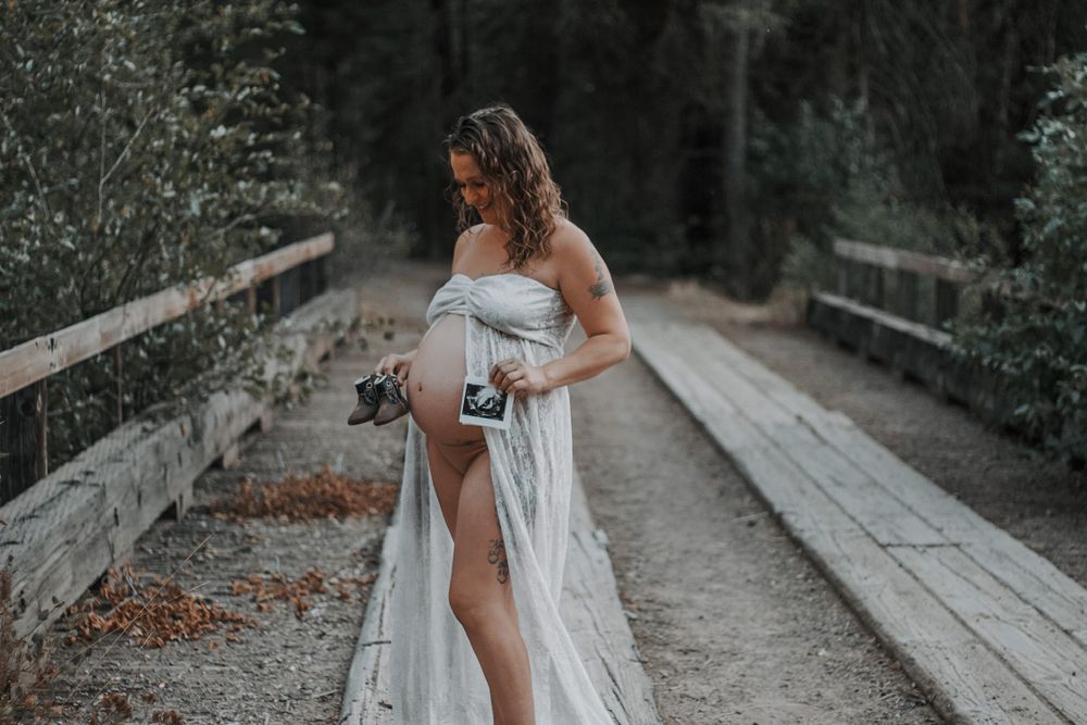 Pregnant woman in white lace wrap holding ultrasound photo on a wooden bridge outdoors.