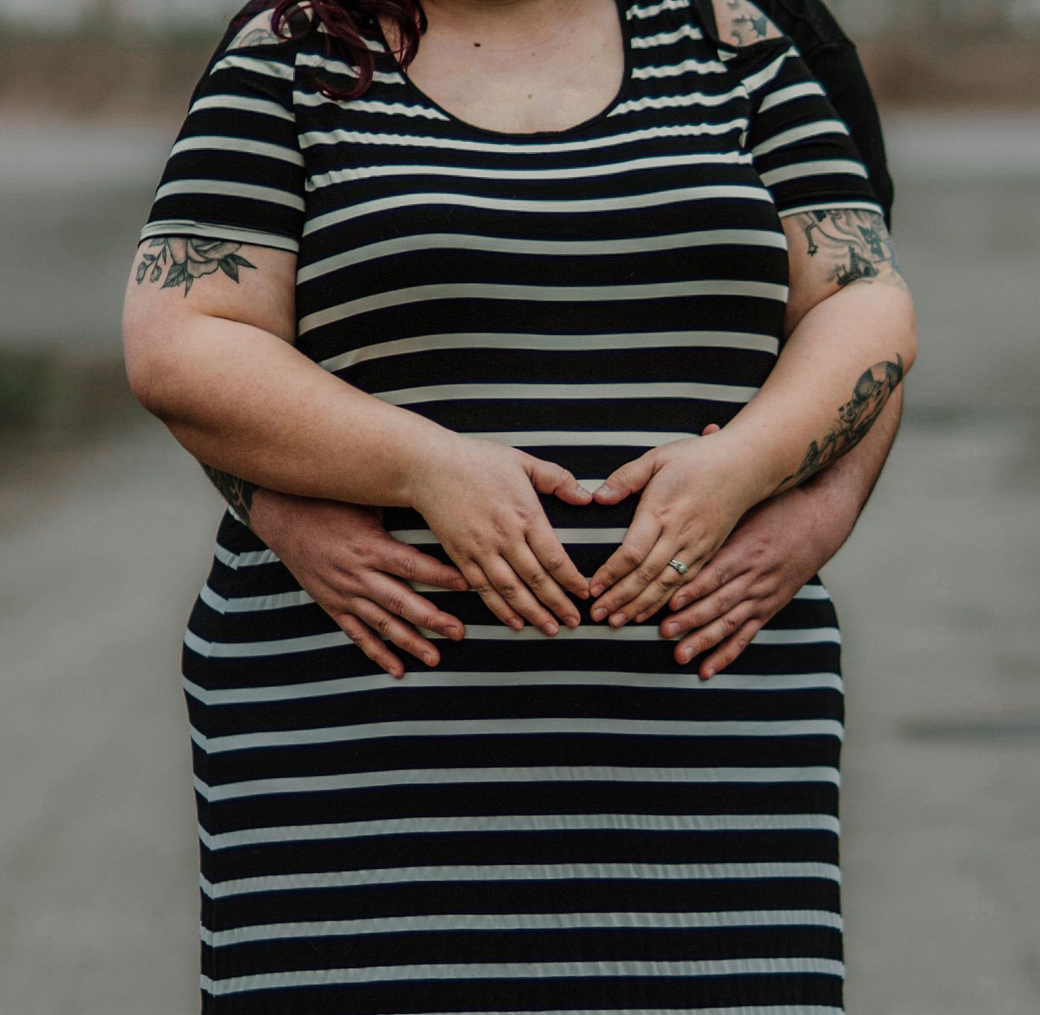 Close-up of couple forming heart shape with hands on pregnant belly in striped maternity dress.