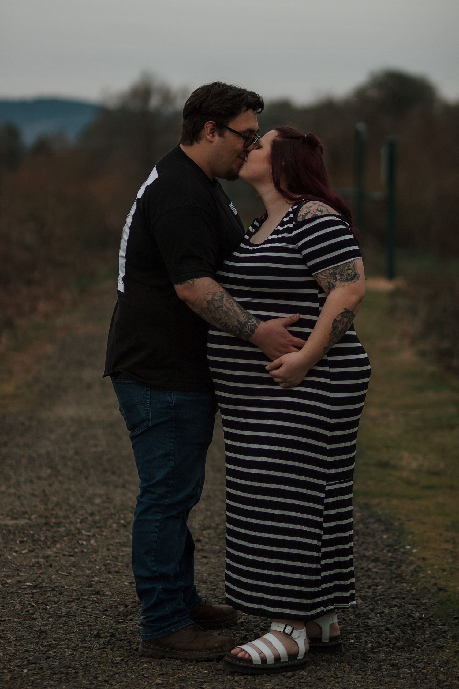 Tattooed couple sharing a kiss outdoors, pregnant woman in striped maxi dress on gravel path.