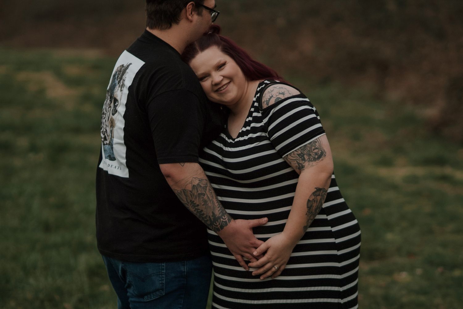 Pregnant woman with tattoos leans on partner while holding hands in lush green outdoor setting.