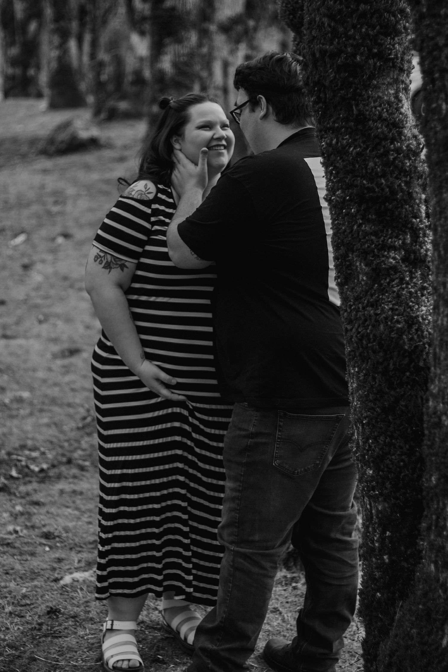 Black and white photo of smiling pregnant woman and partner sharing tender moment by tree.