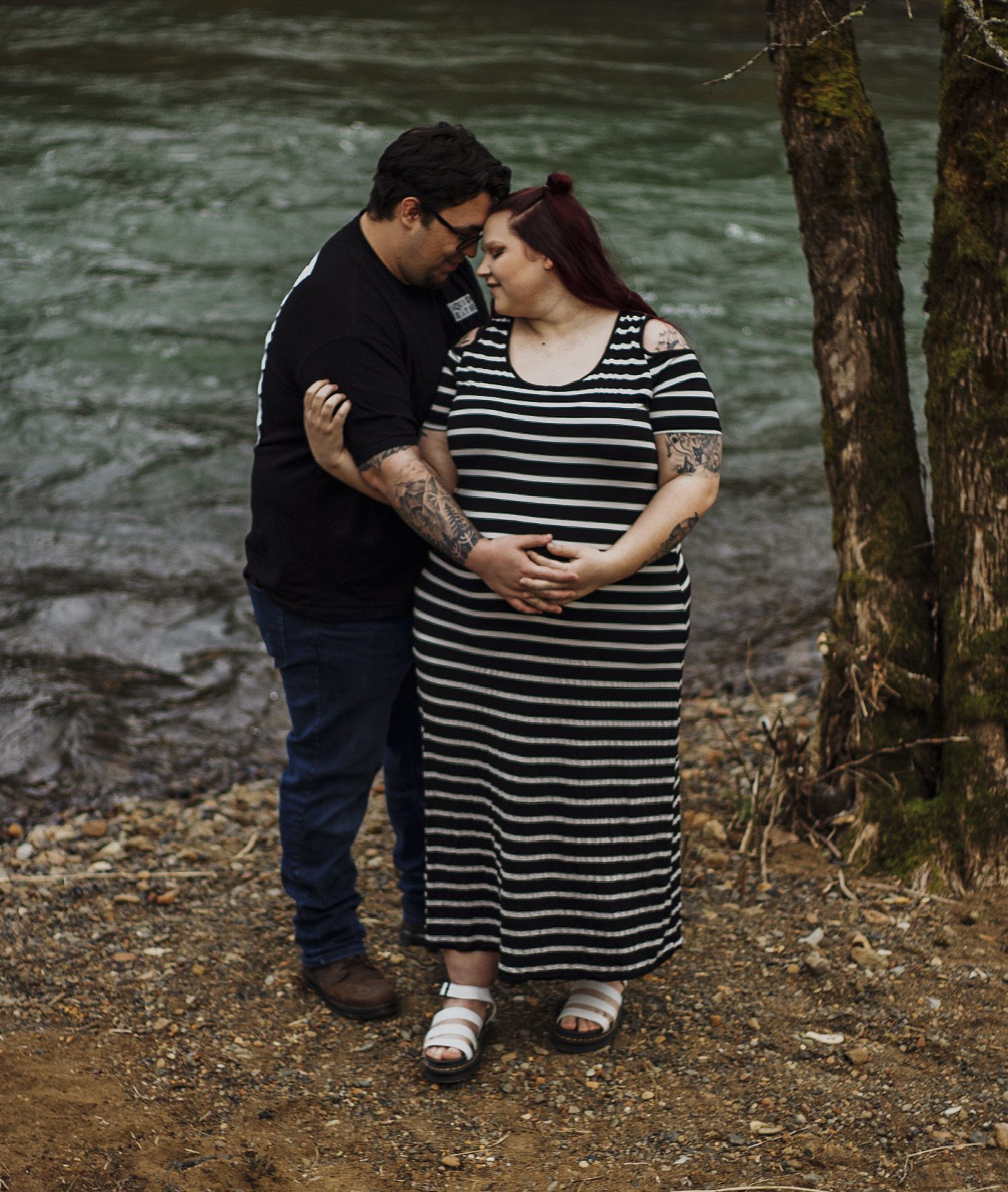 Couple standing by river, hands on pregnant belly, sharing intimate maternity moment.