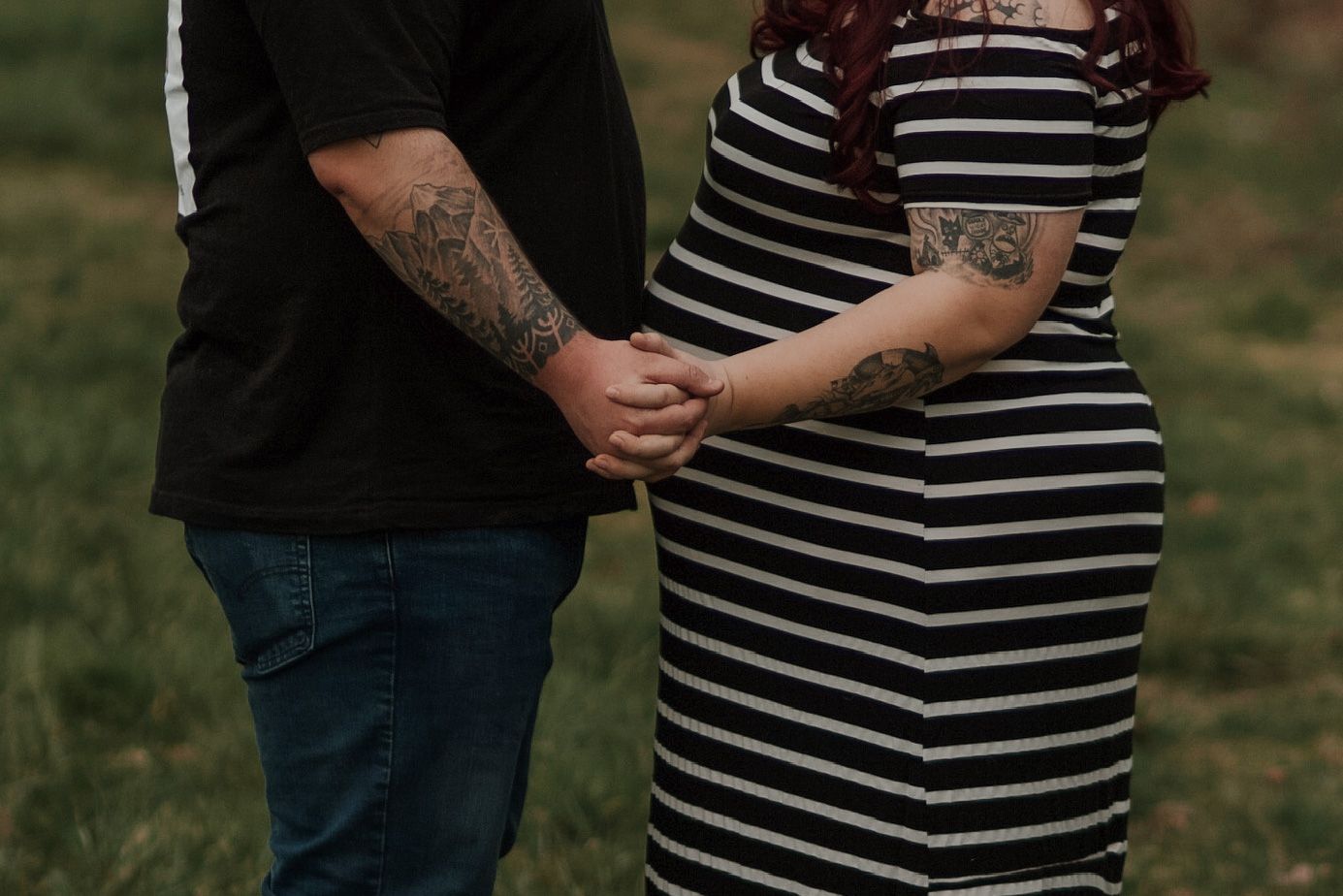 Close-up of tattooed couple holding hands outdoors, pregnant woman wearing black and white striped dress.