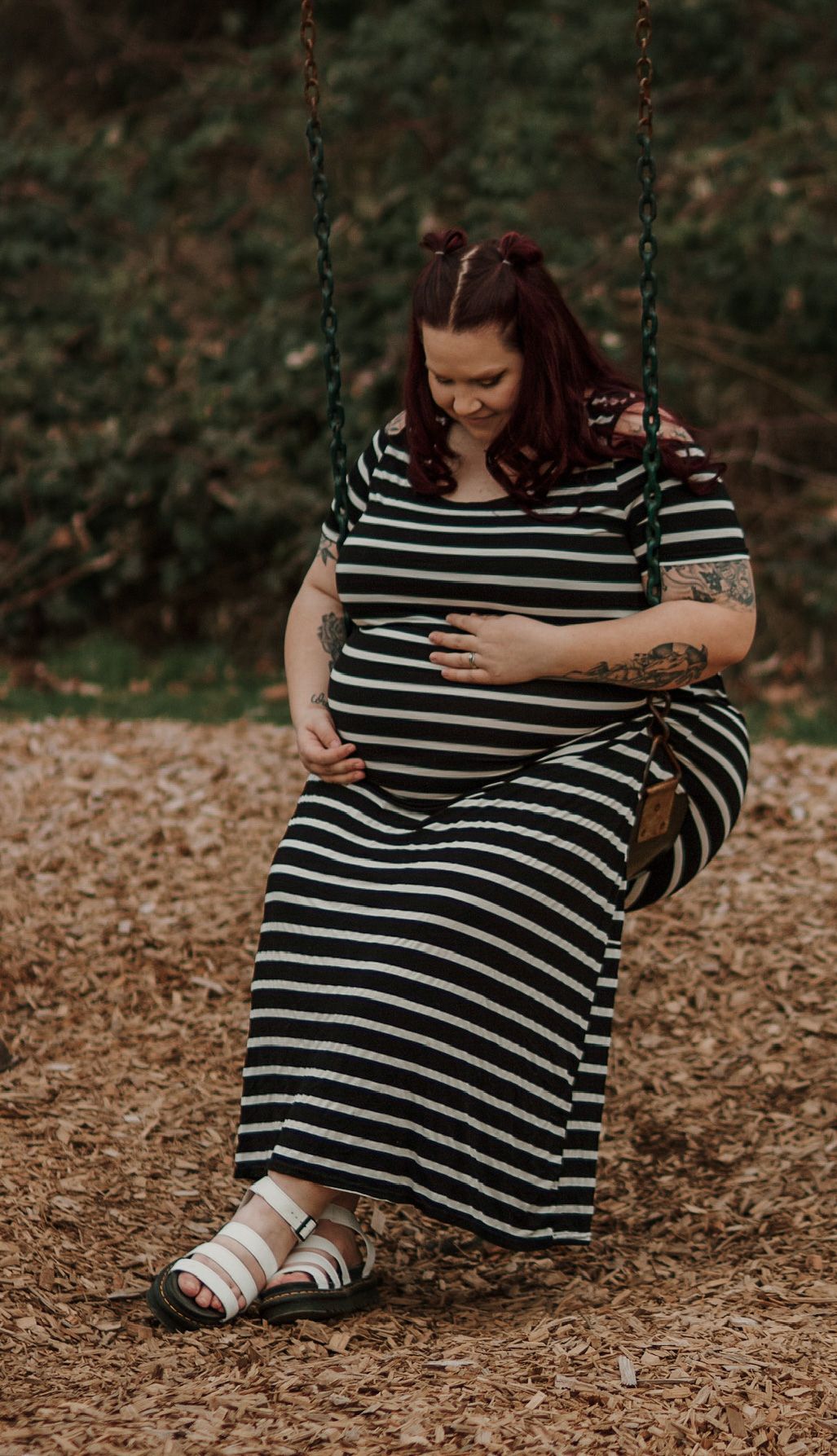Pregnant woman in striped maxi dress sitting on swing, smiling down at baby bump outdoors.