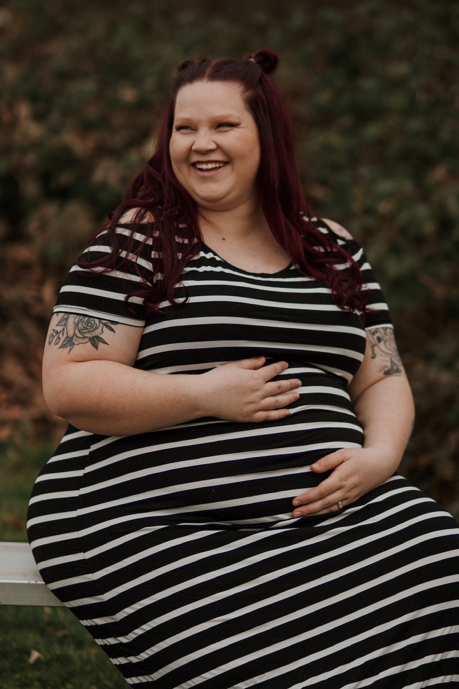 Smiling pregnant woman with red hair and tattoos in striped dress cradling baby bump outdoors.