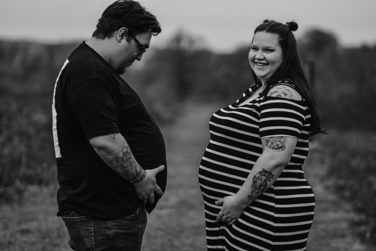 Black and white portrait of tattooed couple facing each other on rural path, woman visibly pregnant.