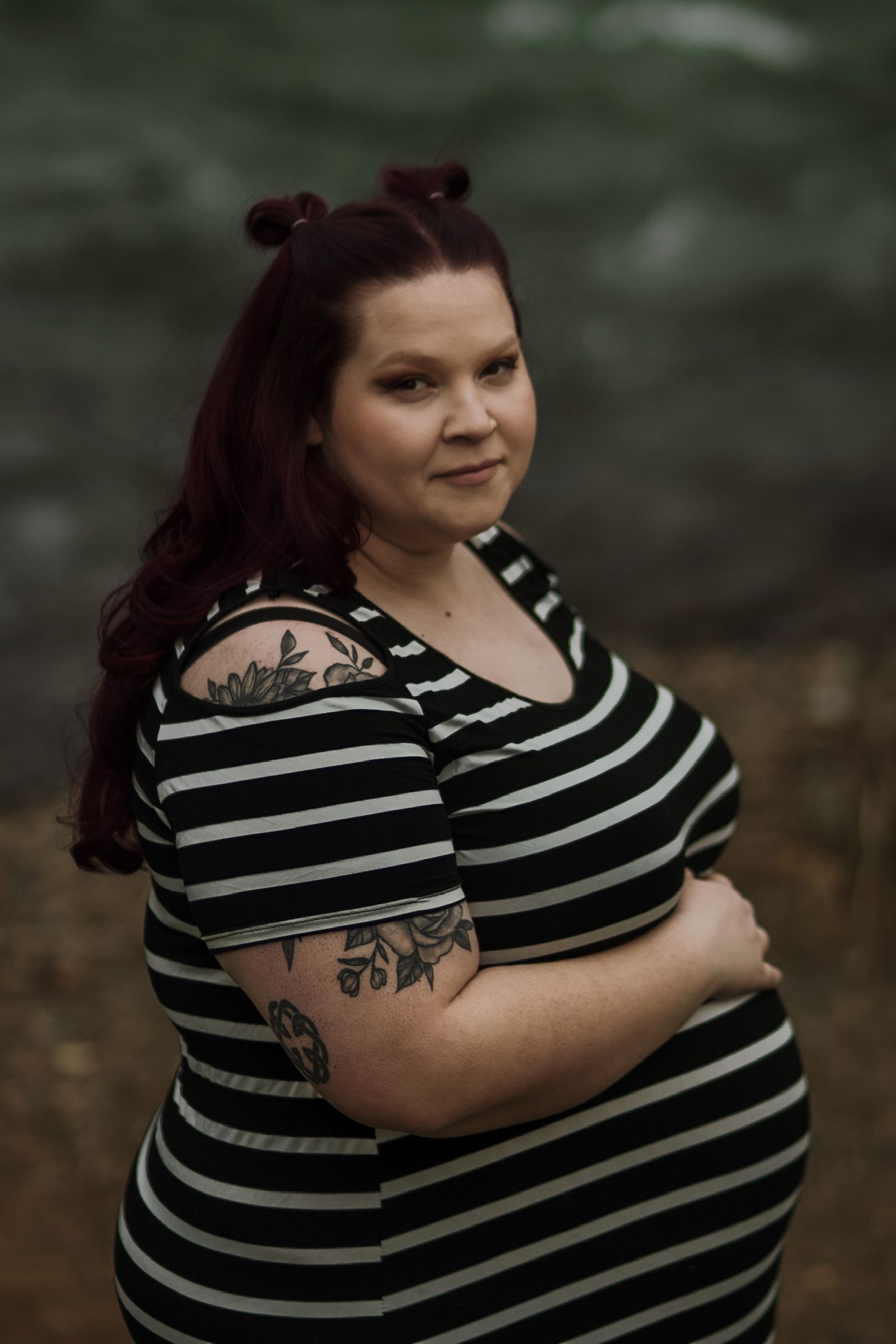 Tattooed pregnant woman in striped dress with red hair posing confidently near water.