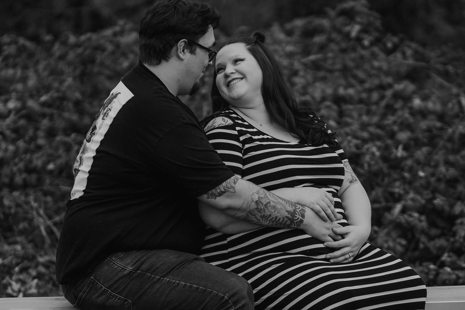 Intimate black and white photo of tattooed couple sharing tender moment, pregnant woman smiling joyfully.