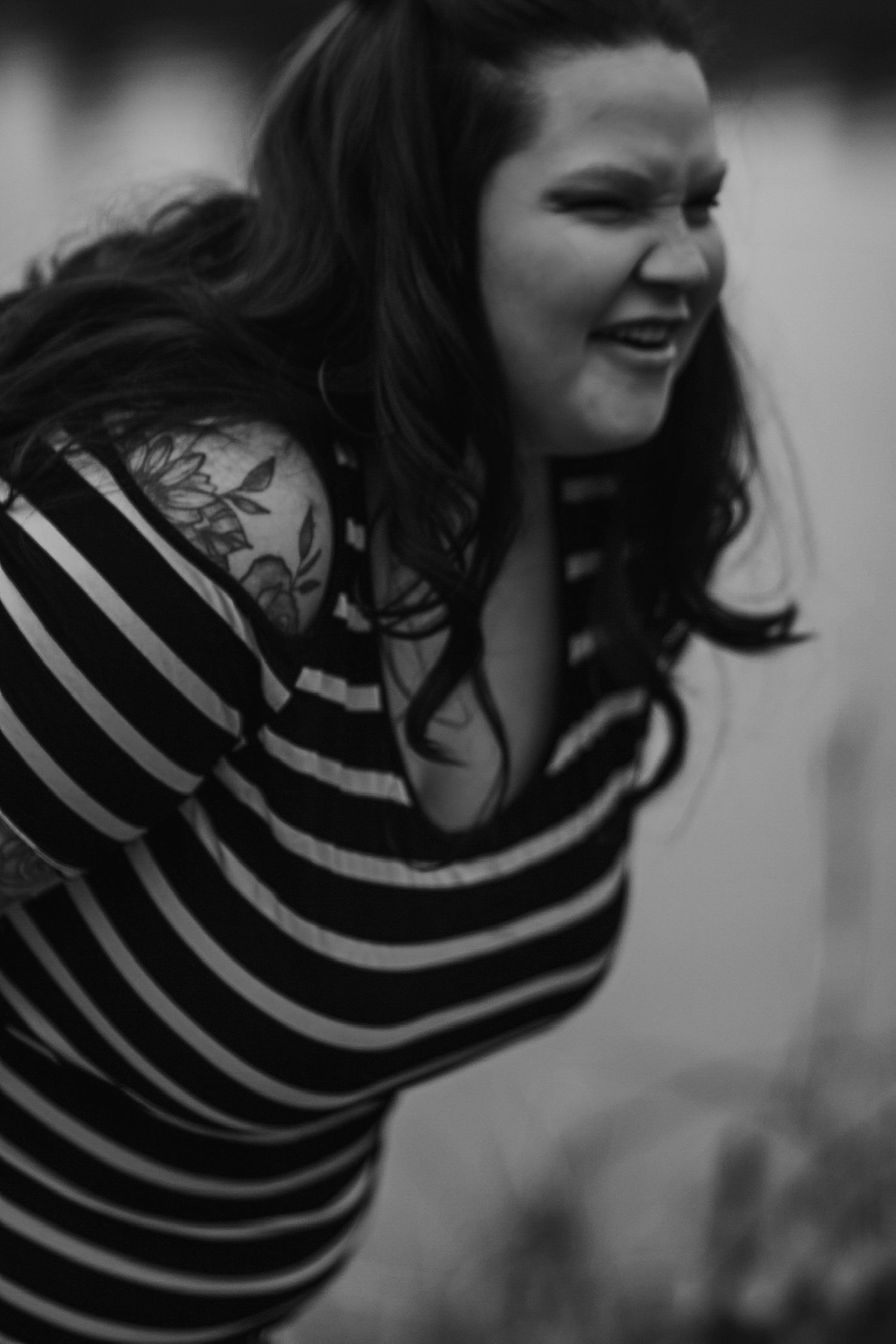 Black and white portrait of laughing pregnant woman with tattoos and striped dress outdoors.