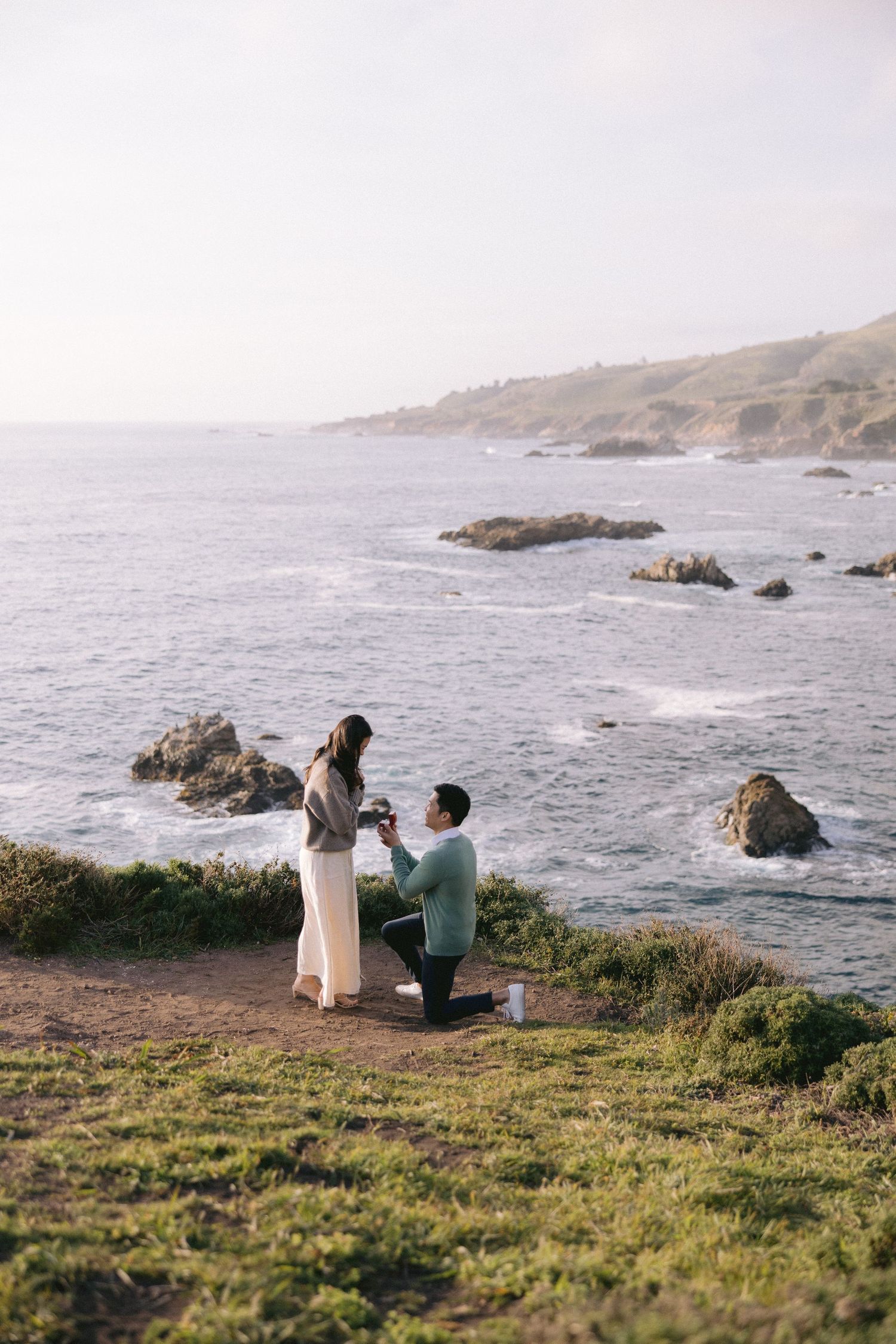 A couple shares a romantic proposal moment on a scenic coastal cliff overlooking the Pacific Ocean during sunset.