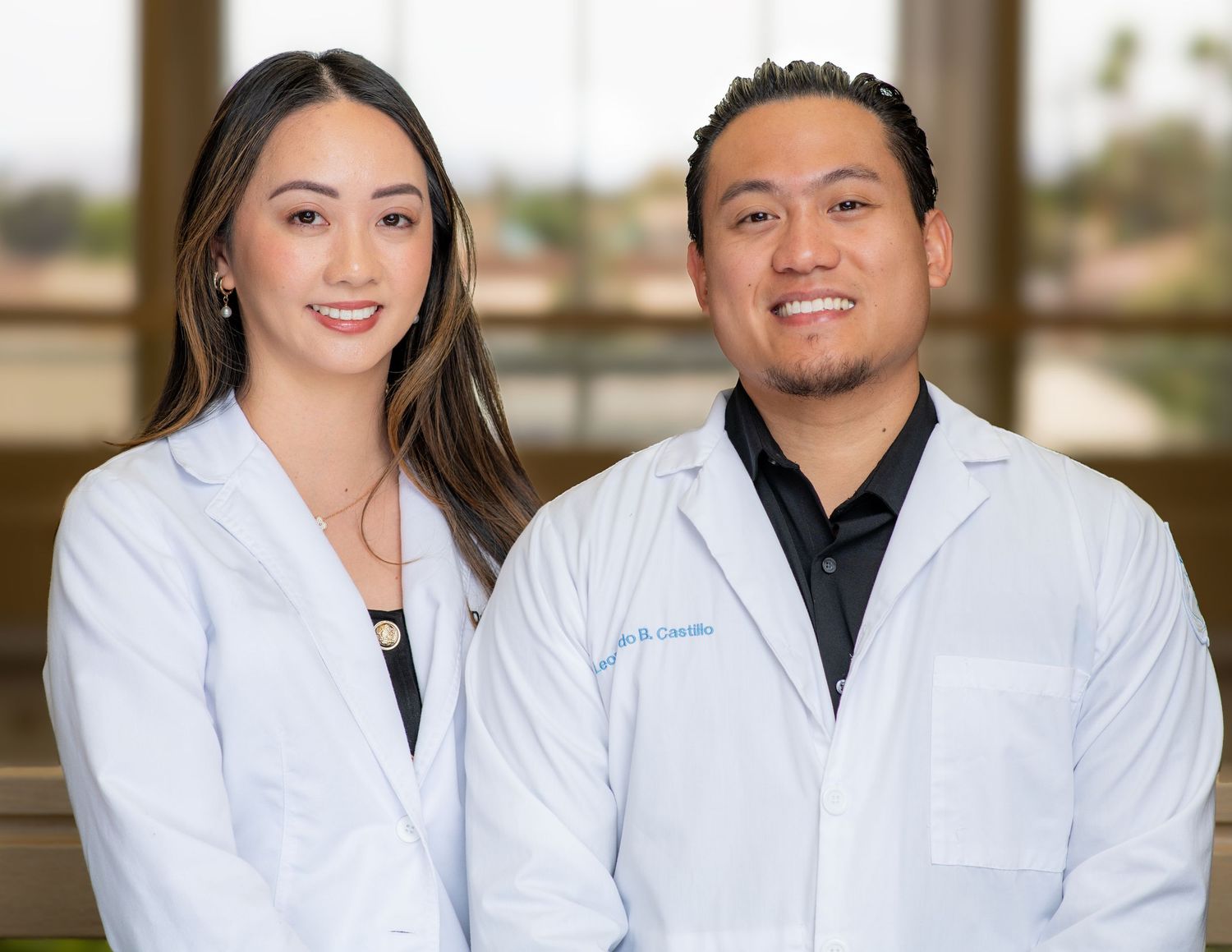 Two healthcare professionals in white lab coats stand together smiling in a medical office setting.