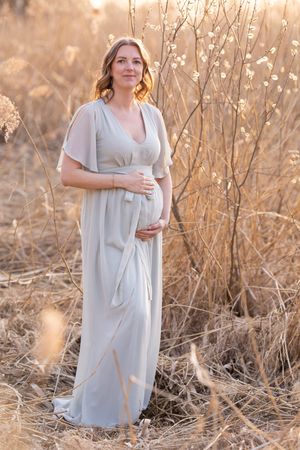 Gravid kvinna i lång ljus klänning på äng i kvällsol, gravidfotografering Västerås, MemoriesbyM