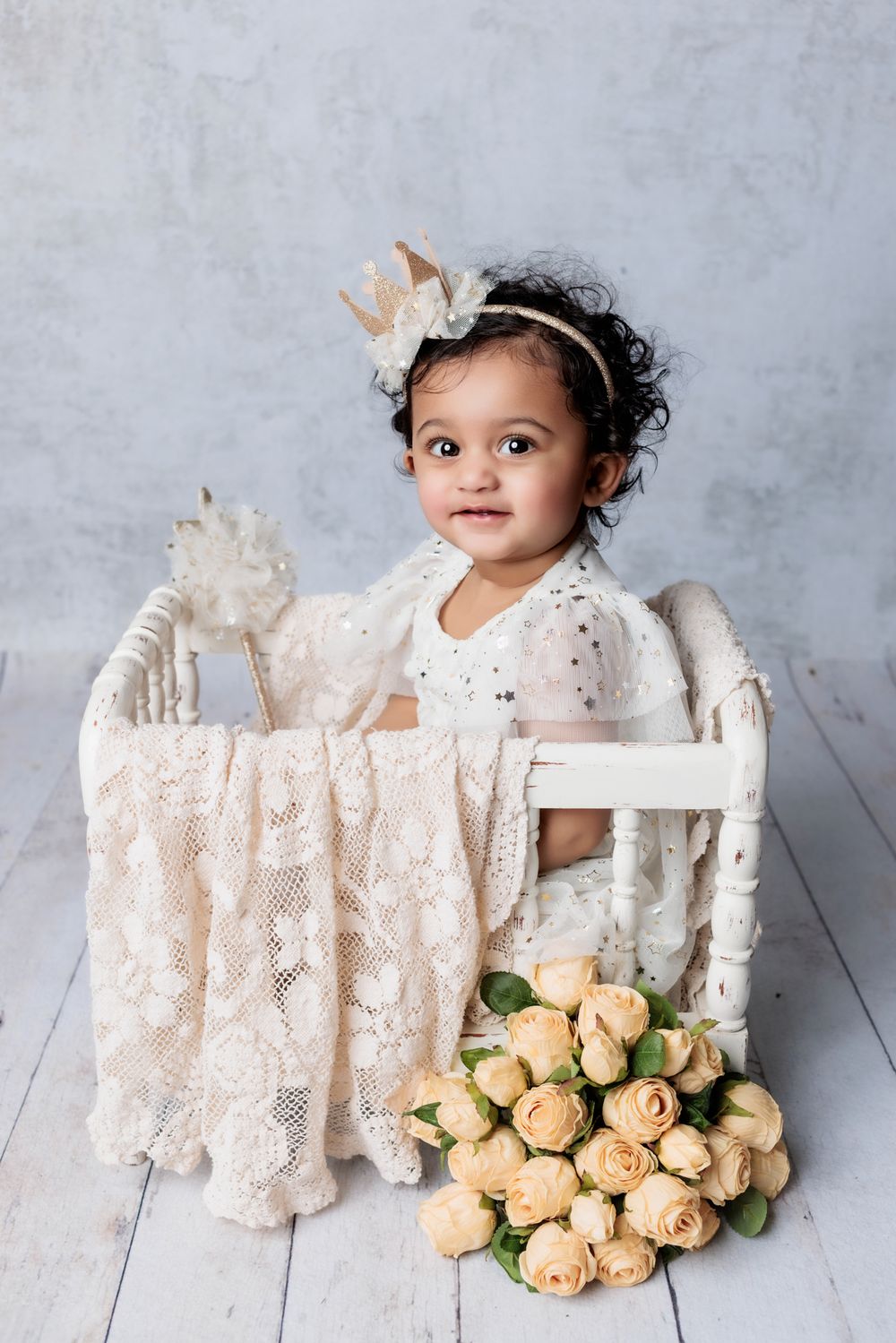 A baby wearing a white lace dress sits in a vintage white crate beside peach roses on a light wooden floor.