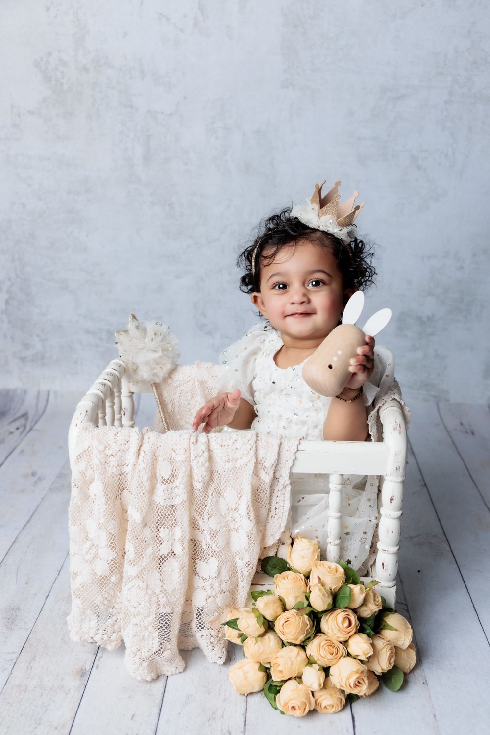 A studio portrait with a child seated in a white wooden chair with cream lace fabric and peach roses nearby.