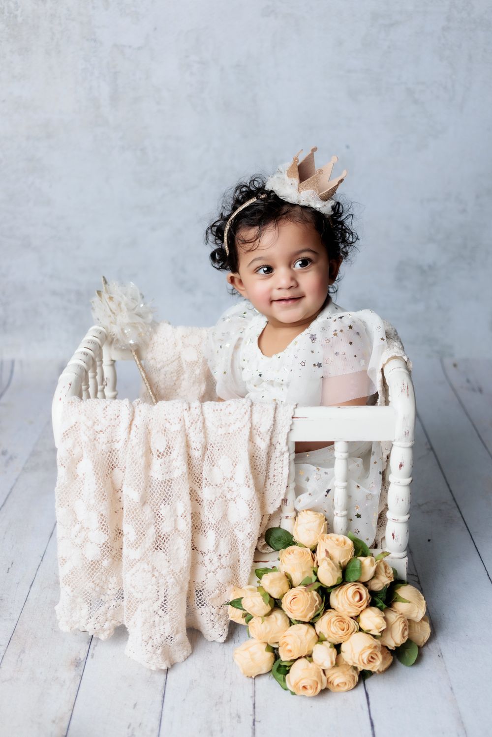 Small child in white dress sitting in vintage white chair with pink roses and lace decor against grey backdrop.