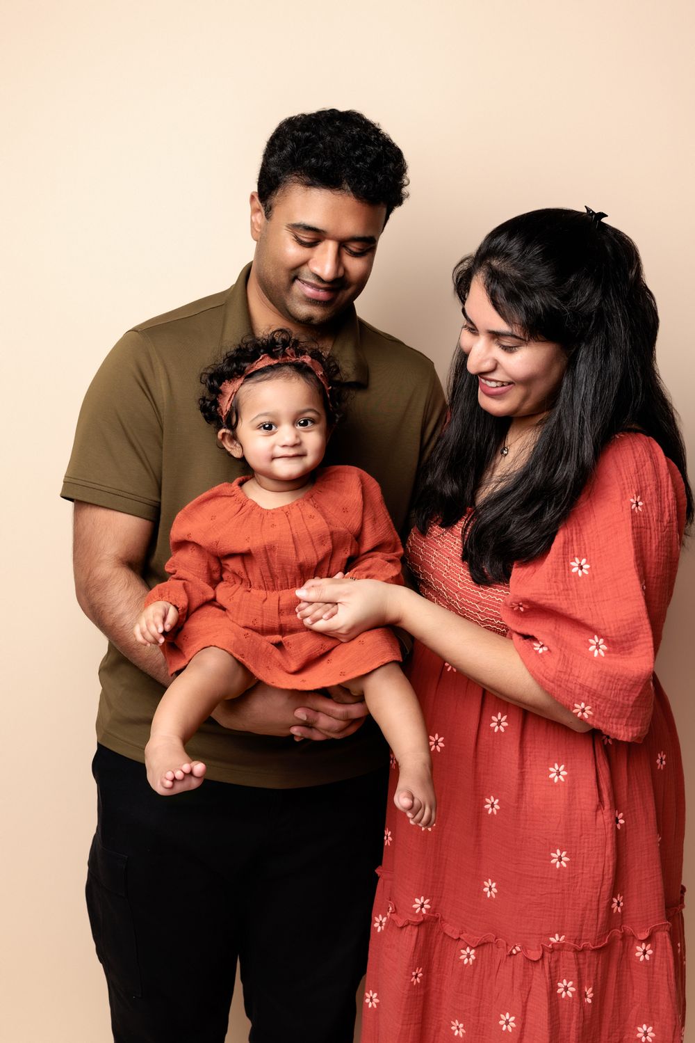 A loving family portrait showing parents in earth tones and red floral dress holding their smiling baby in a coral outfit.