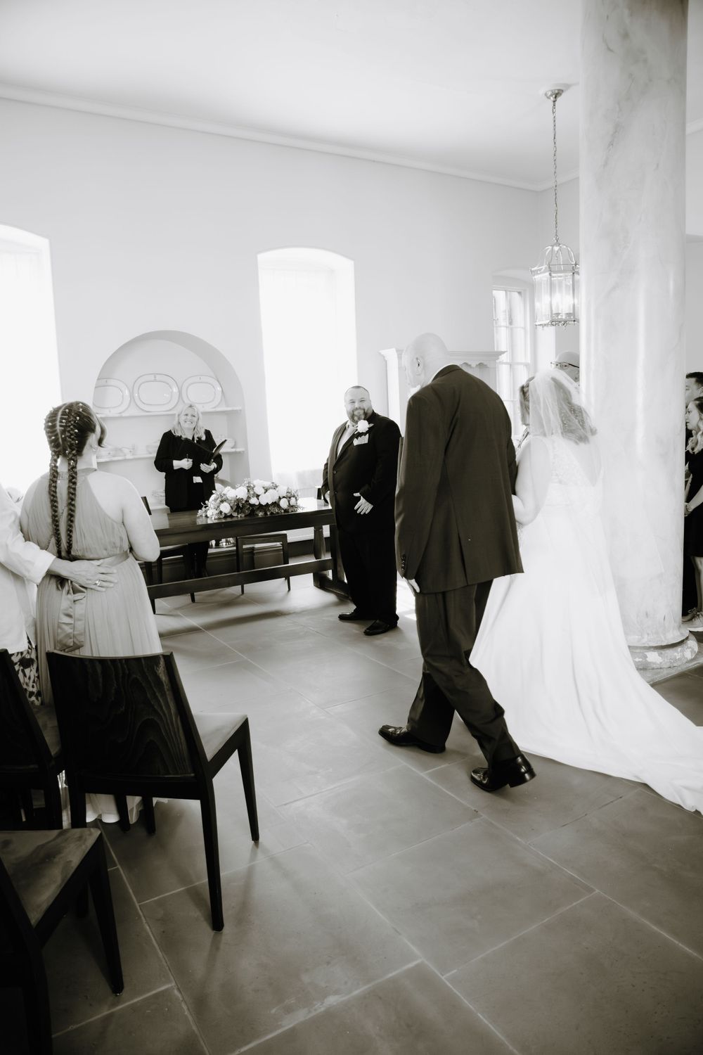 A black and white photo of wedding guests in formal attire gathered inside an elegant room with arched windows.