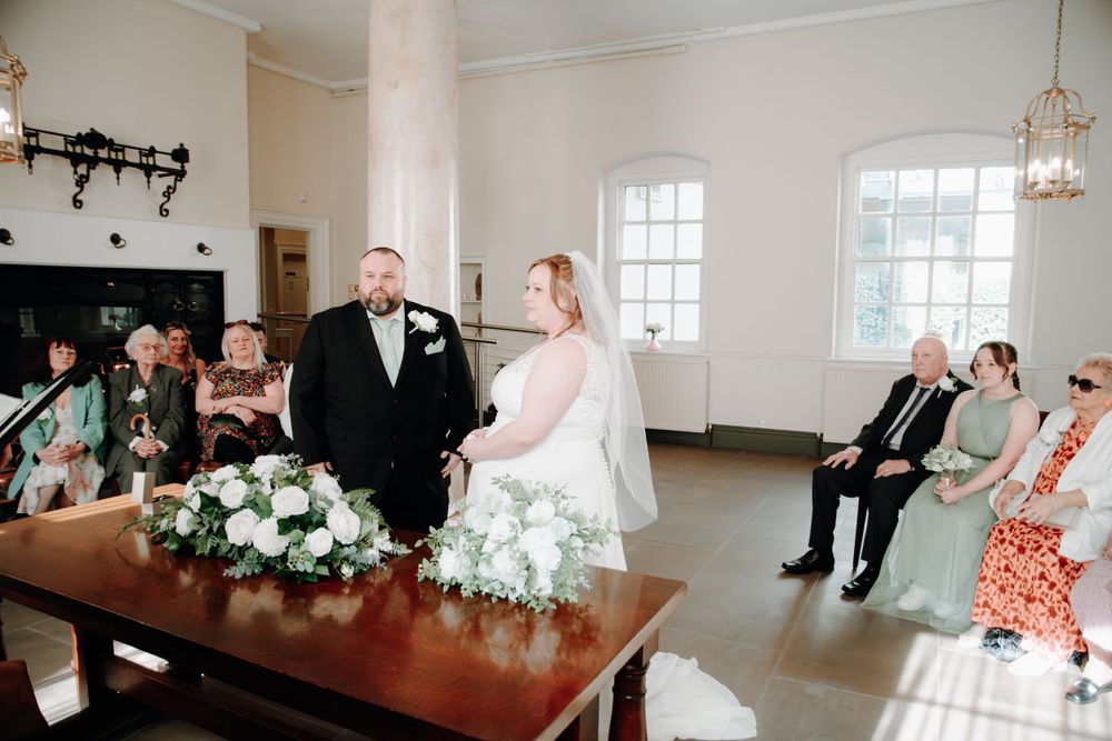 Indoor wedding ceremony in a bright room with white floral arrangements and guests seated on wooden benches.