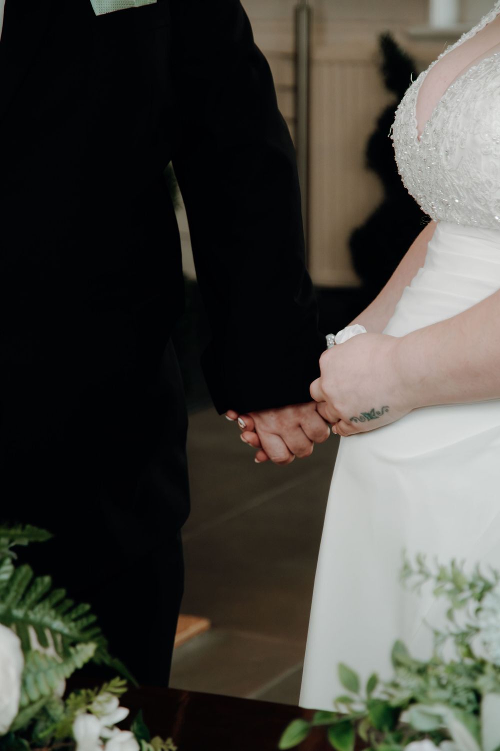 Close-up photo of bride in white dress and groom in black suit holding hands during wedding ceremony with greenery.