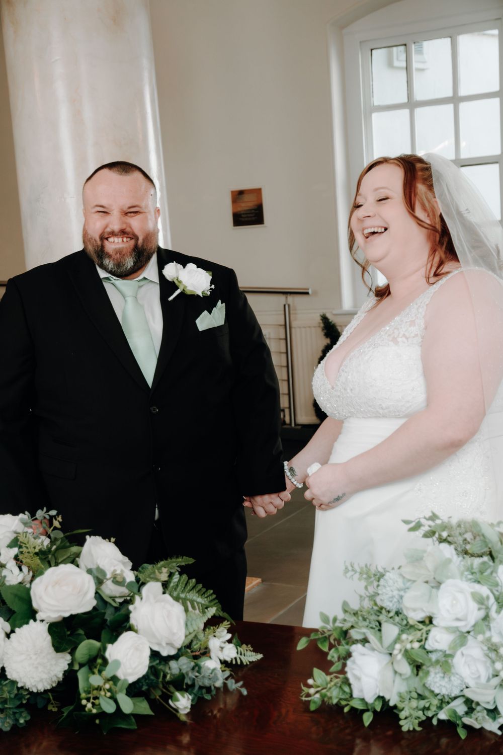 A bride and groom share a joyful moment during their wedding ceremony, with white floral arrangements on the table.