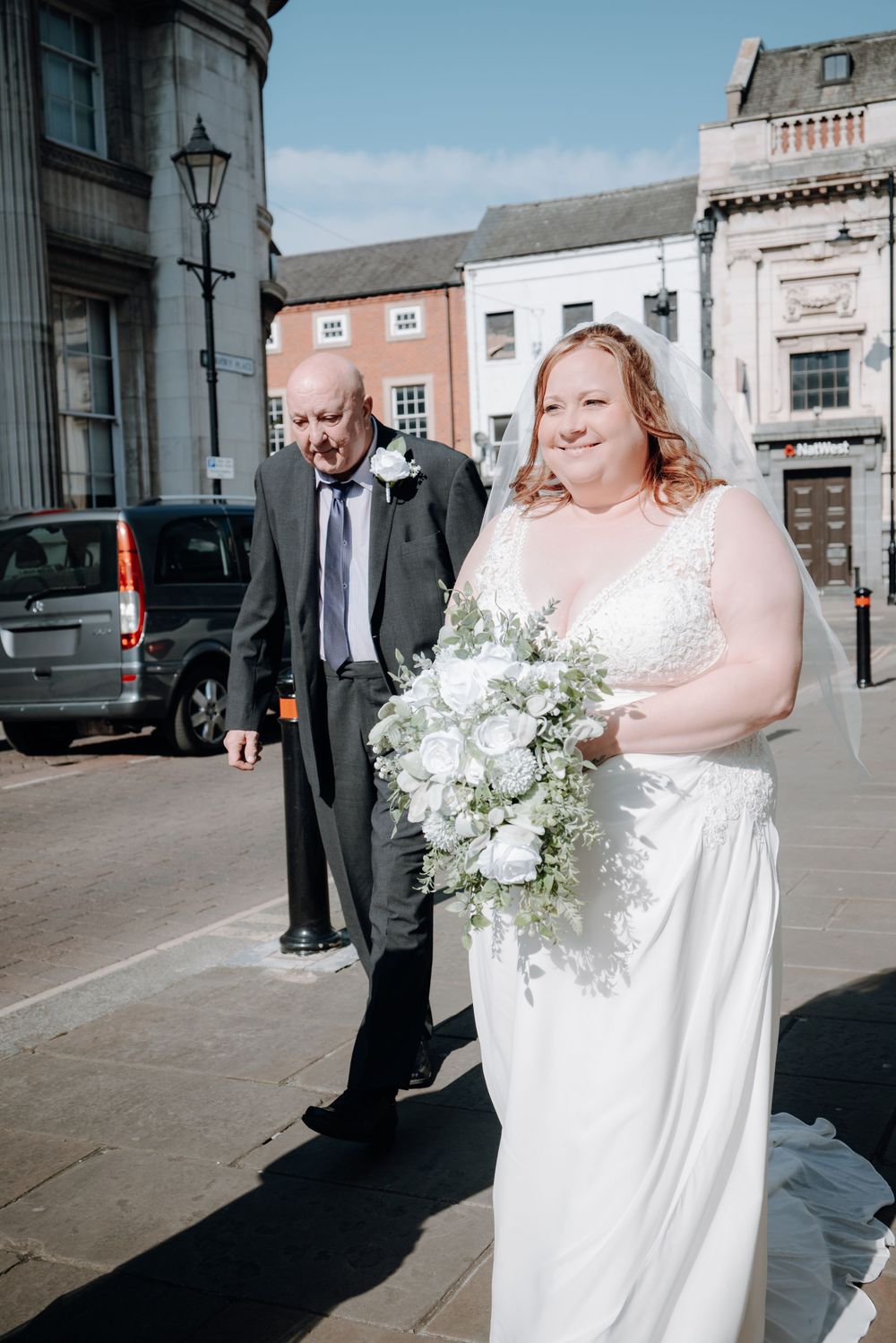 A bride in a white wedding dress holds a beautiful white floral bouquet while standing on a city street.