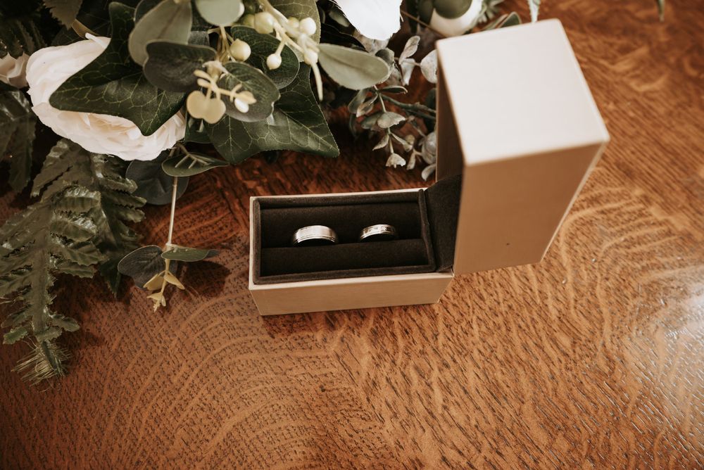 Wedding rings displayed in a black box on a wooden surface with eucalyptus branches and white ribbon decoration.