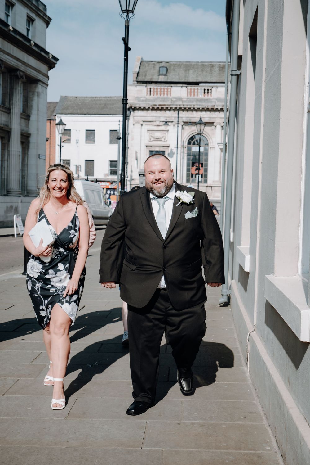 A couple walks down a sunny urban street with white buildings and a vintage lamp post.