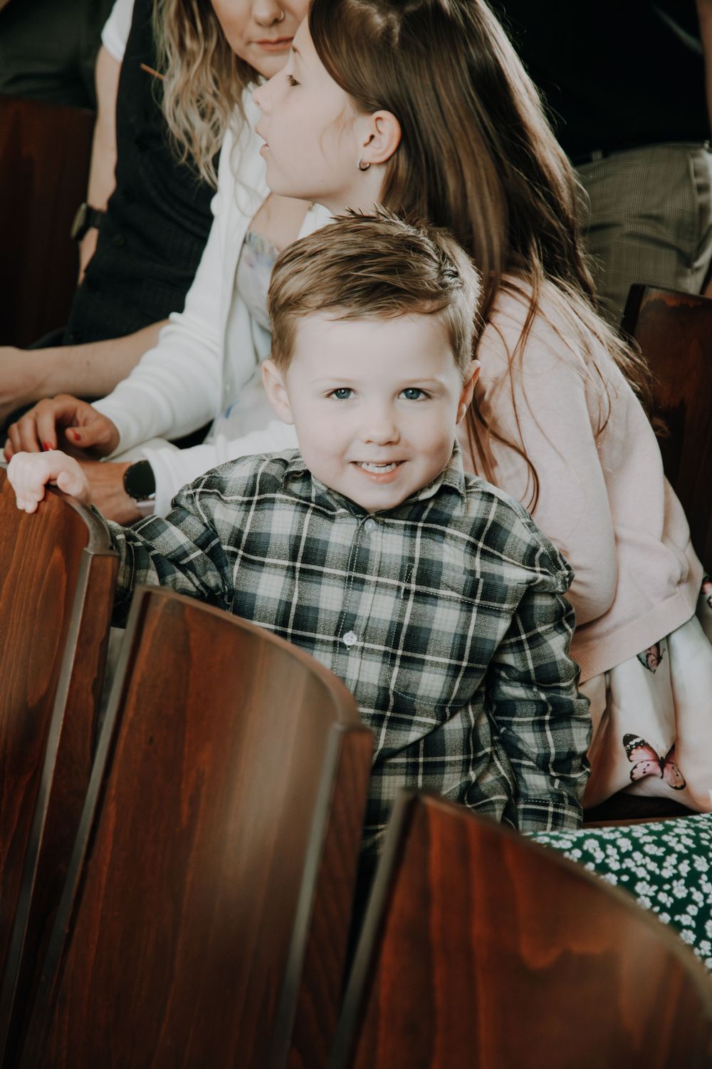 A young child wearing a plaid button-up shirt smiles while sitting in a wooden chair at an indoor gathering.