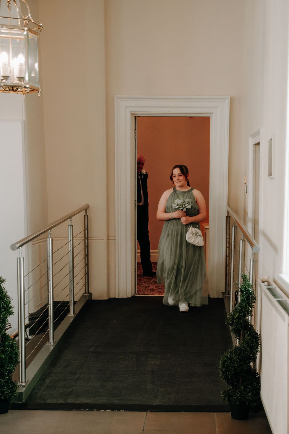A green-dressed figure stands in an elegant hallway with white walls and a metal railing, holding flowers and a small purse.