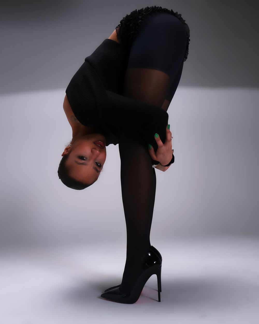 Artistic portrait of person in black attire performing flexible pose in stiletto heels against grey background.