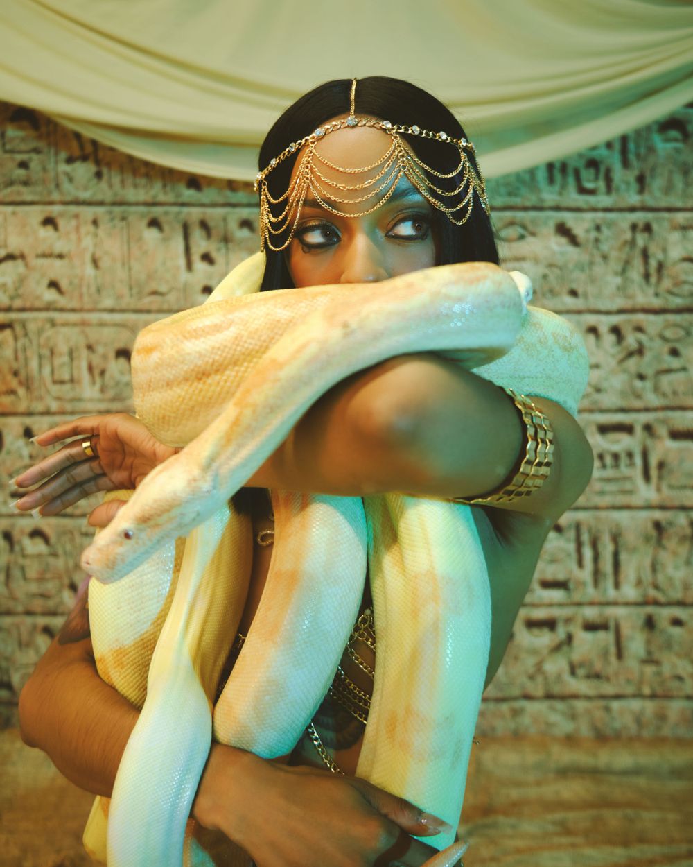 Fashion portrait with golden headpiece and flowing fabric against hieroglyphic wall.