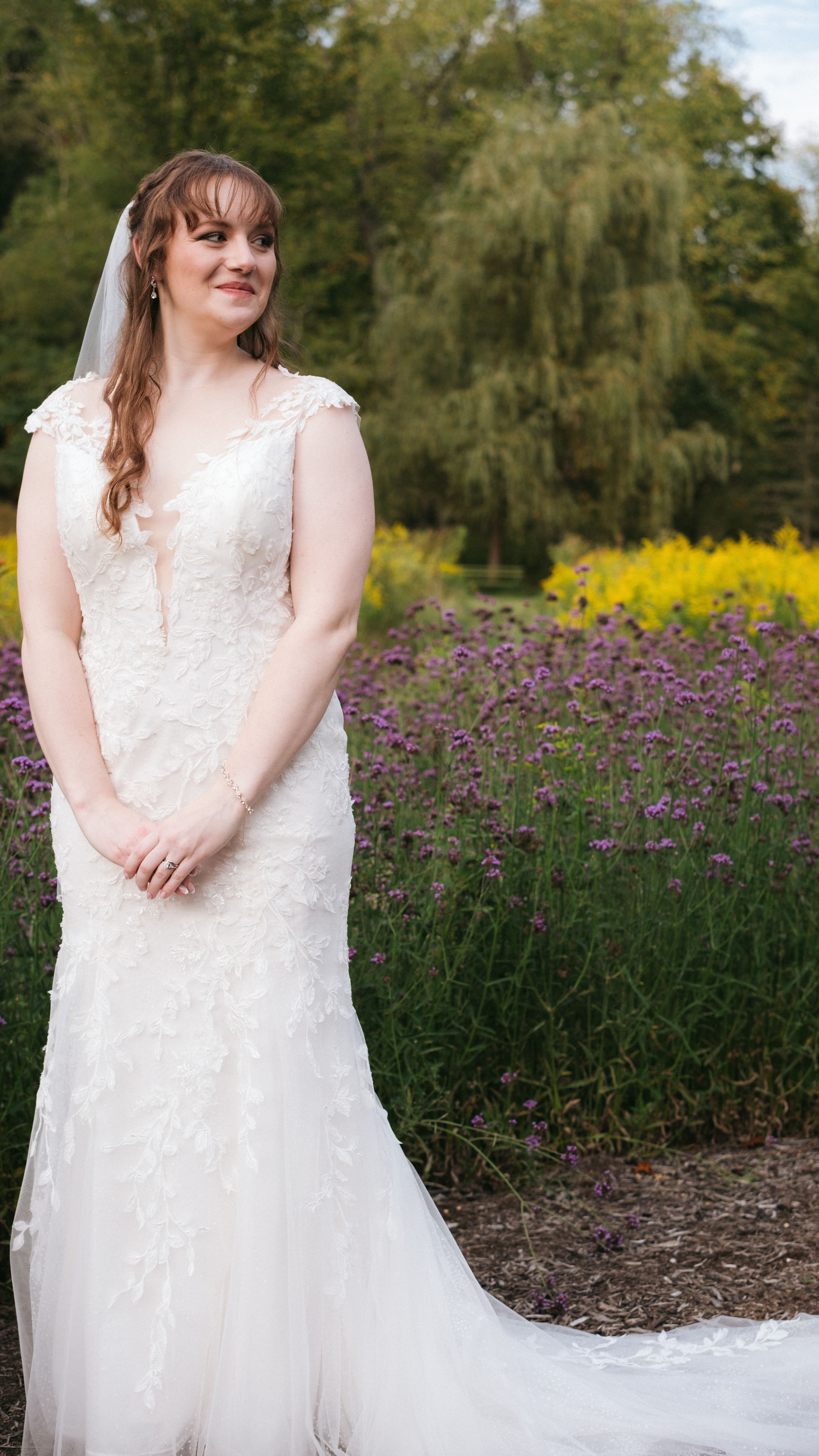 A bride in a white lace gown stands in a garden with purple wildflowers and yellow blooms in the background.