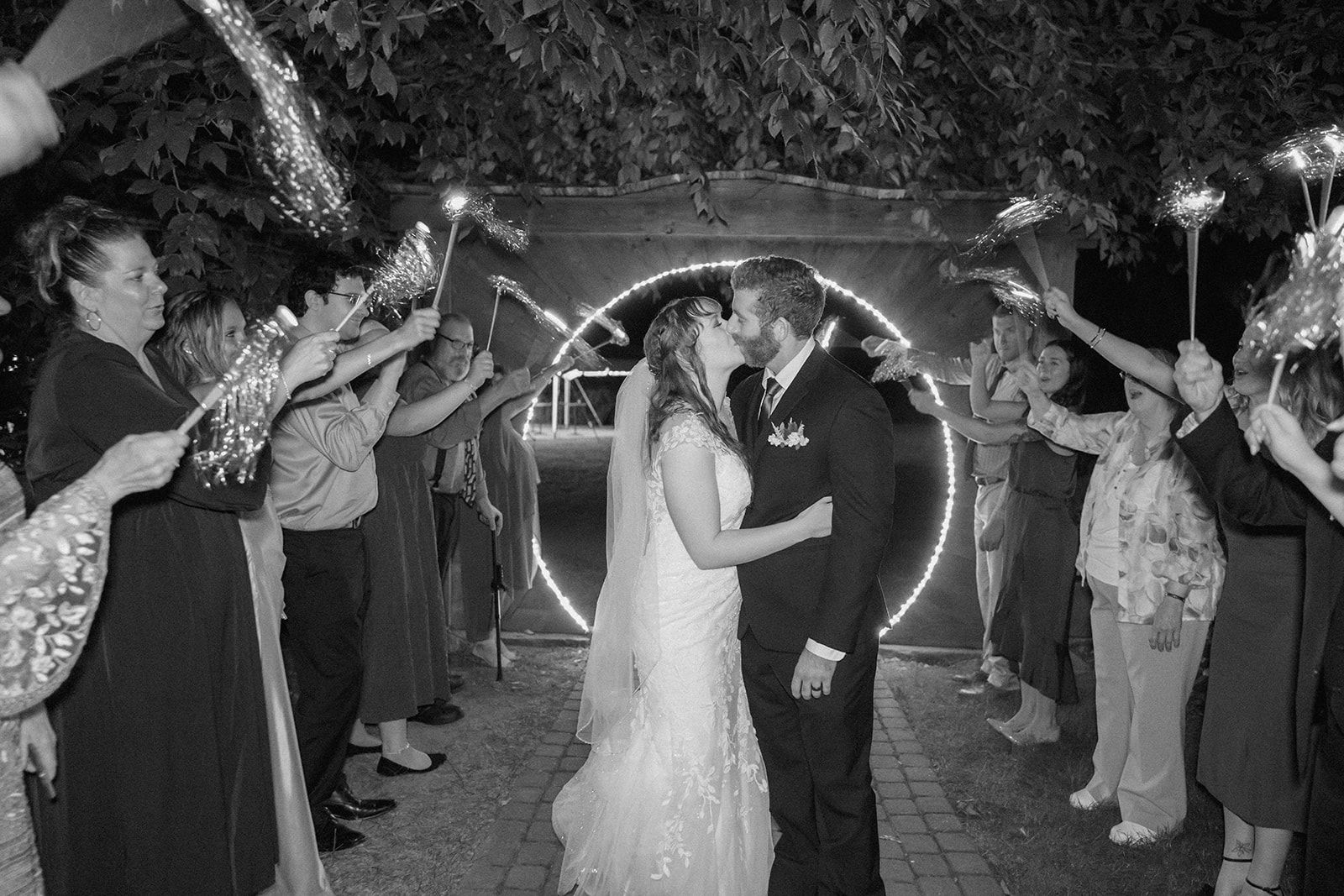 Wedding guests hold sparklers forming a glowing archway as newlyweds share a romantic moment in black and white photography.
