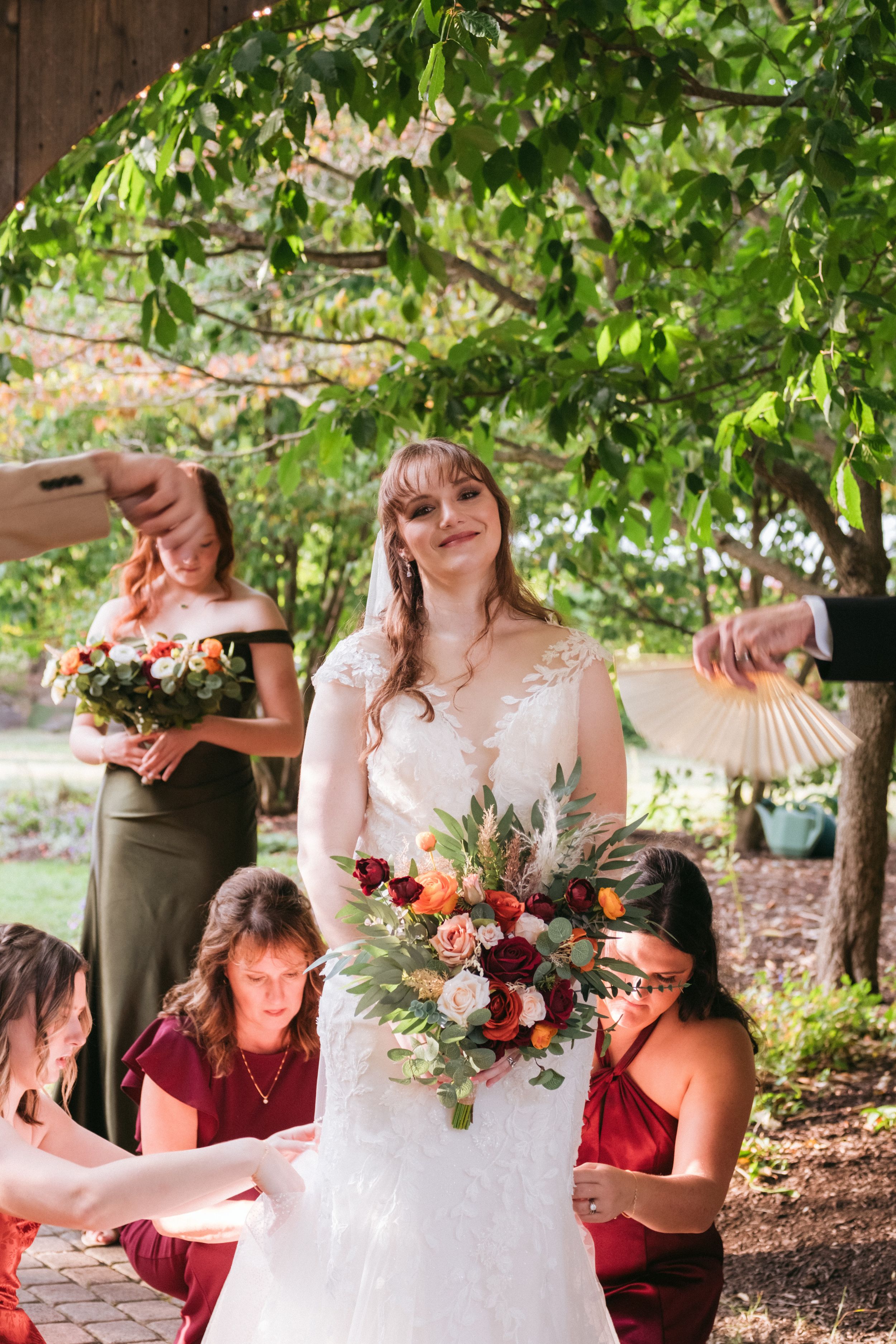 A person in a white lace wedding dress holds a colorful bouquet with burgundy flowers while standing outdoors under trees.