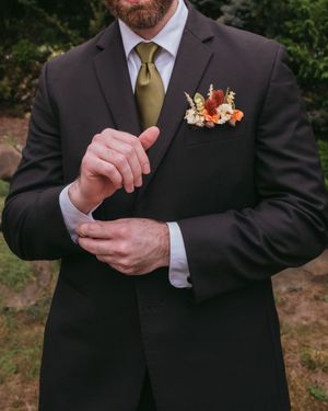 Close-up of person in dark suit adjusting cufflink with autumn-colored floral boutonniere on lapel.