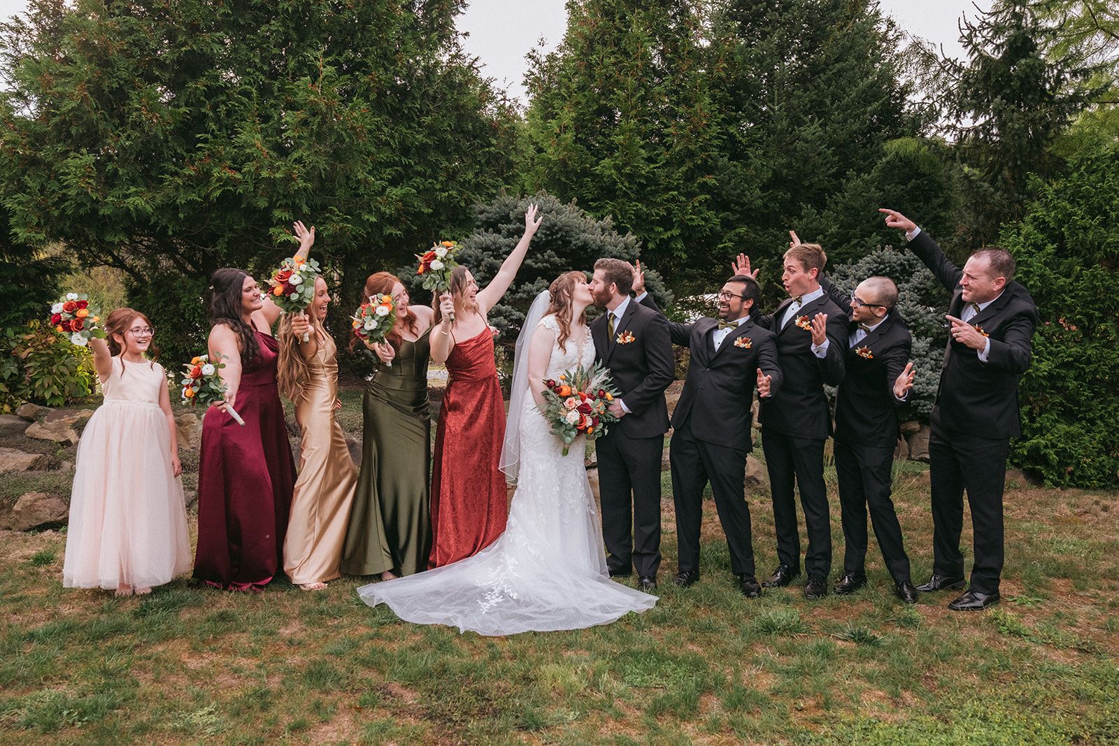 A wedding party poses together outdoors with bridesmaids in mixed-color dresses and groomsmen in dark suits.