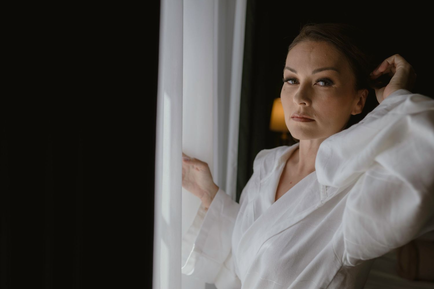 Artistic portrait of person in white robe looking out window during wedding preparations.