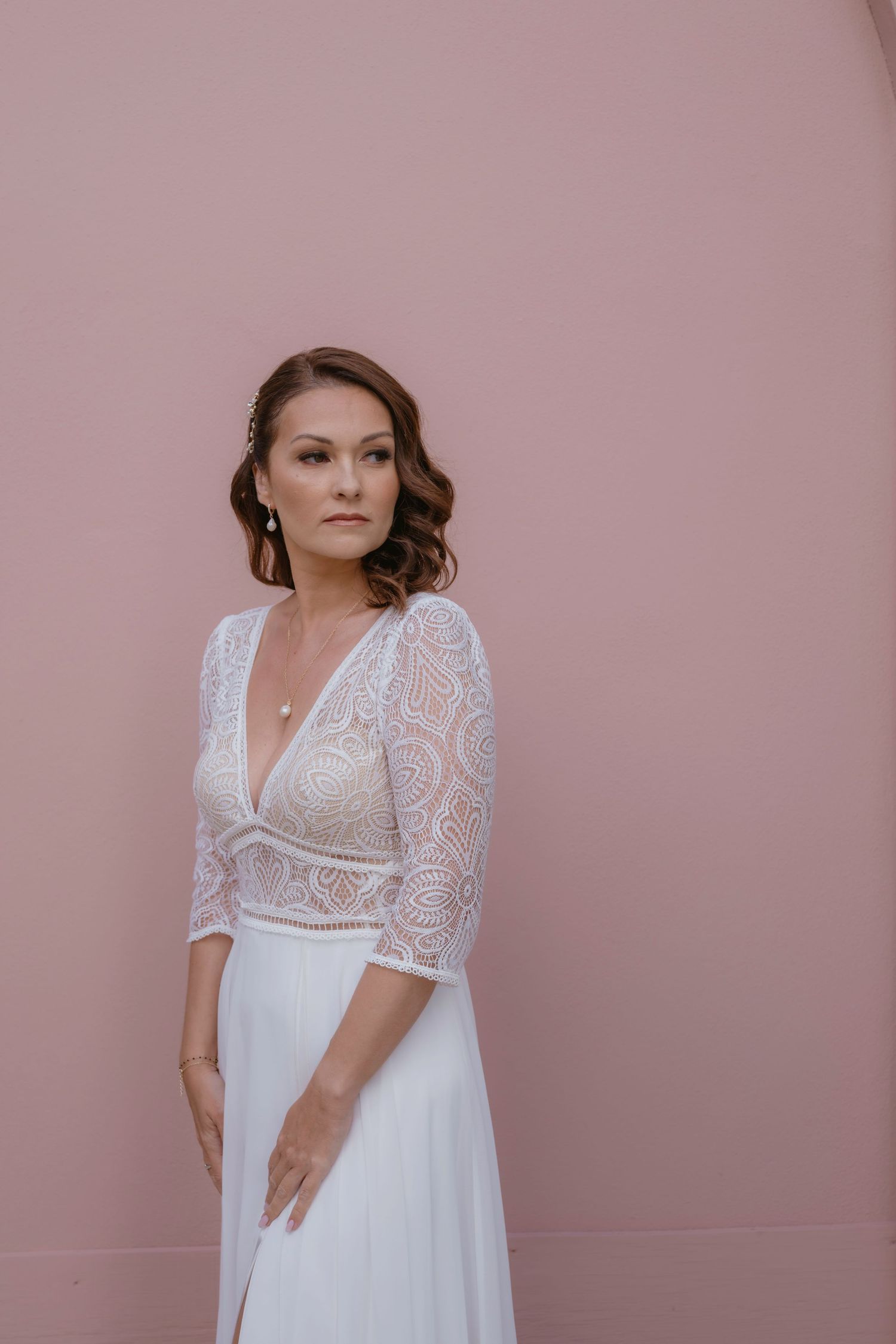 Bride in elegant lace-sleeved wedding dress poses against pink wall.