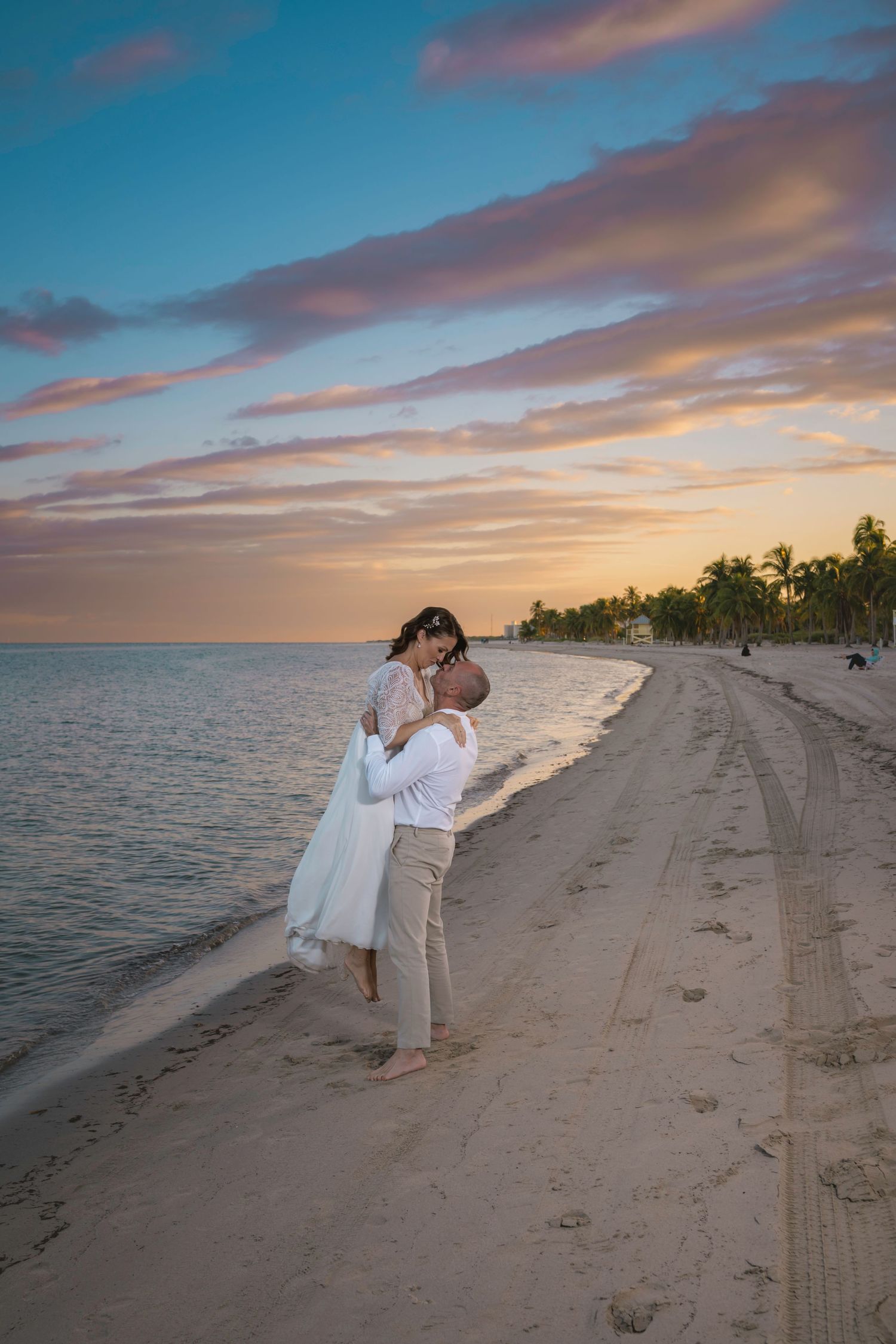 A romantic beach wedding photo of a couple embracing during golden hour with palm trees in the distance.