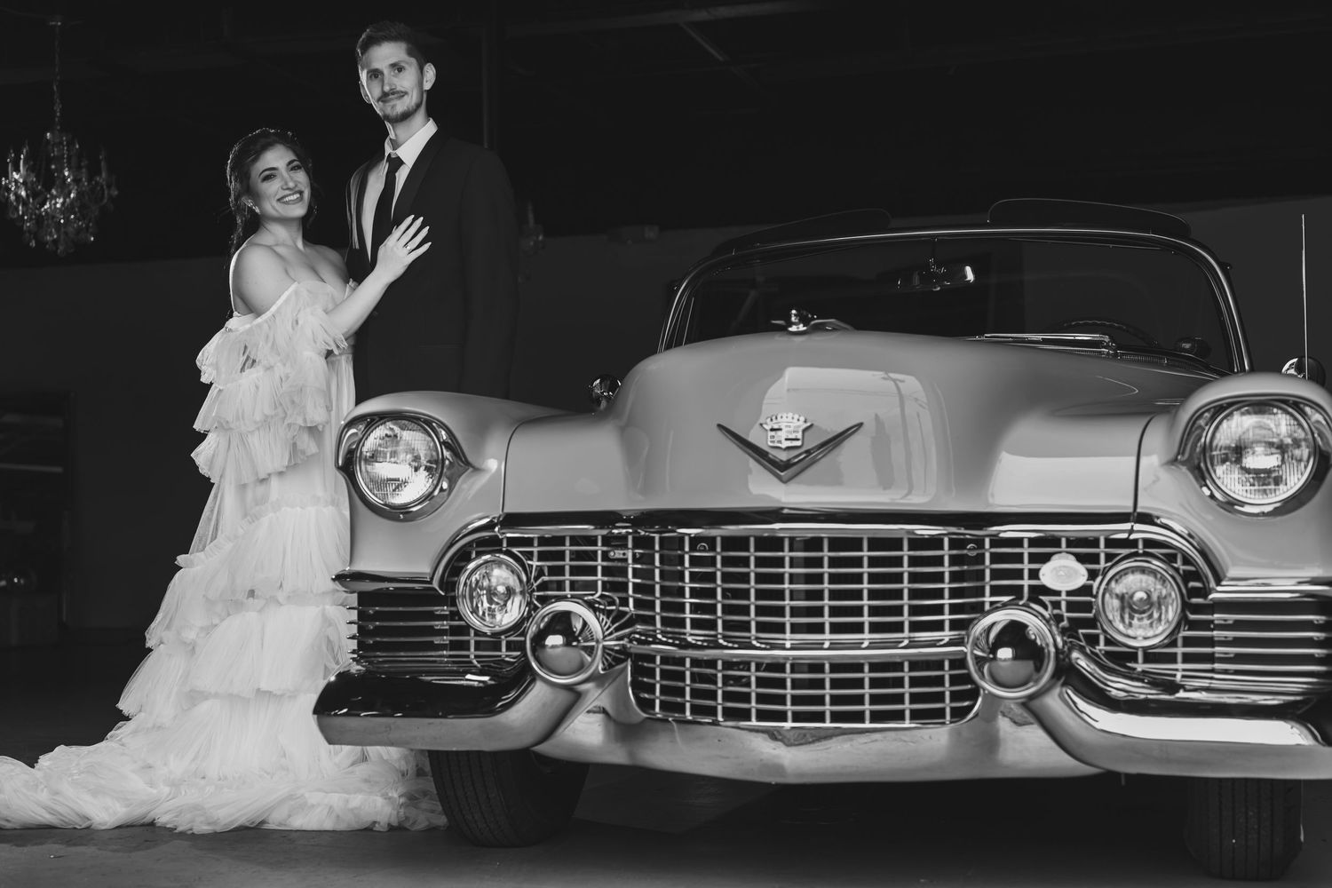 A black and white portrait of newlyweds posing with a classic Cadillac.
