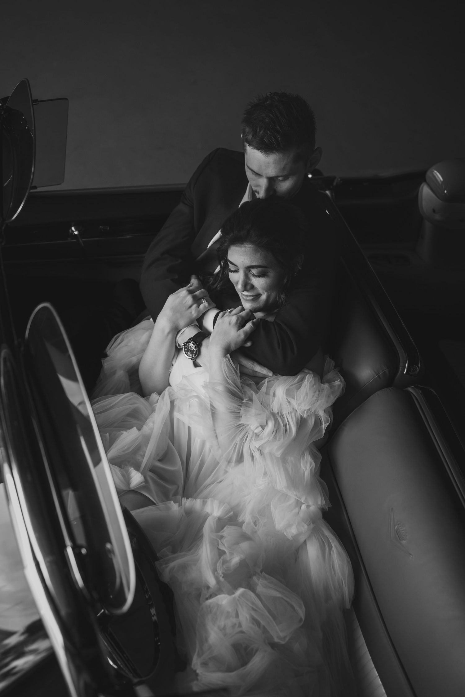 Romantic black and white portrait in classic car interior with flowing white dress details.