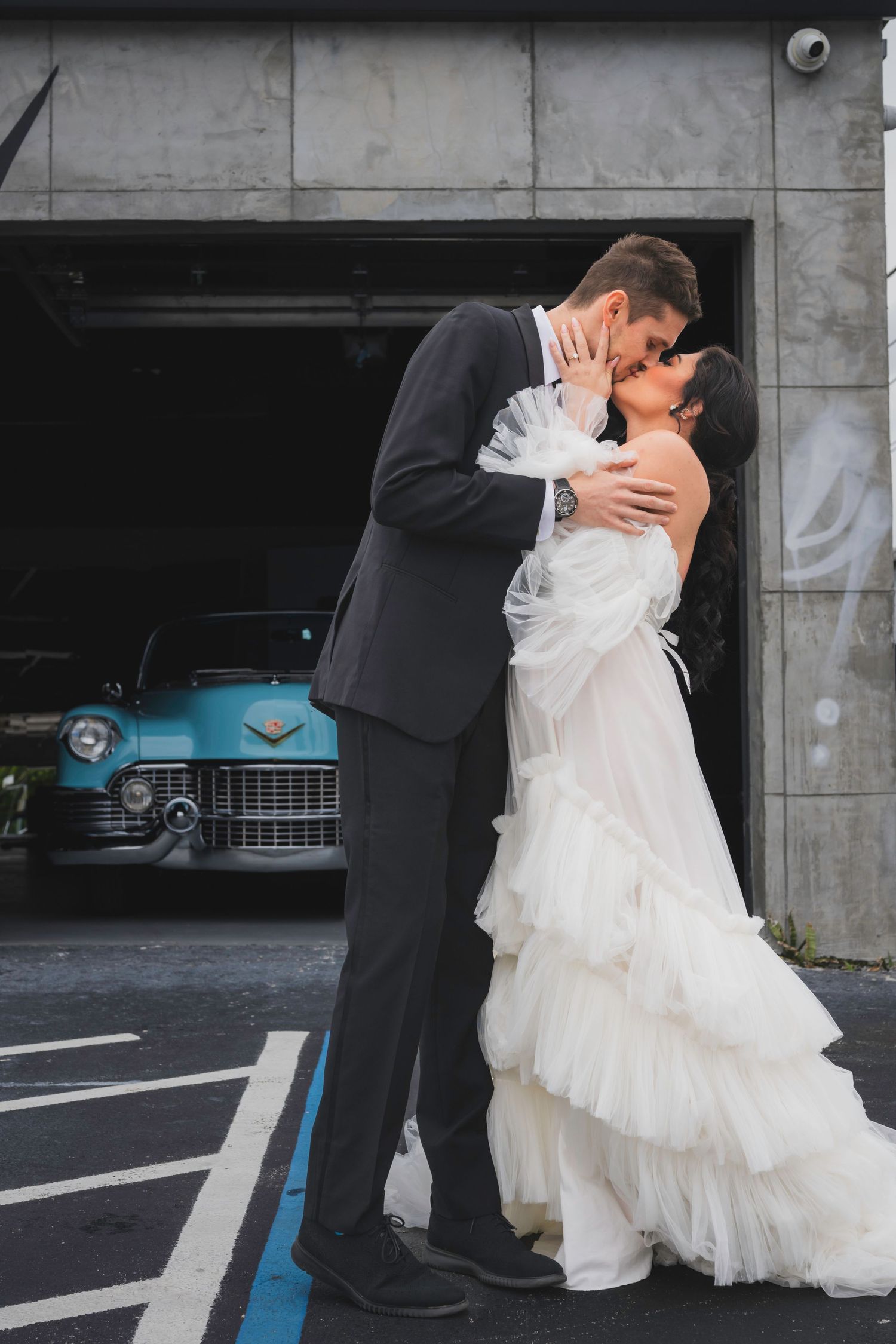 Newlywed couple shares romantic kiss in parking garage with vintage blue car in background.