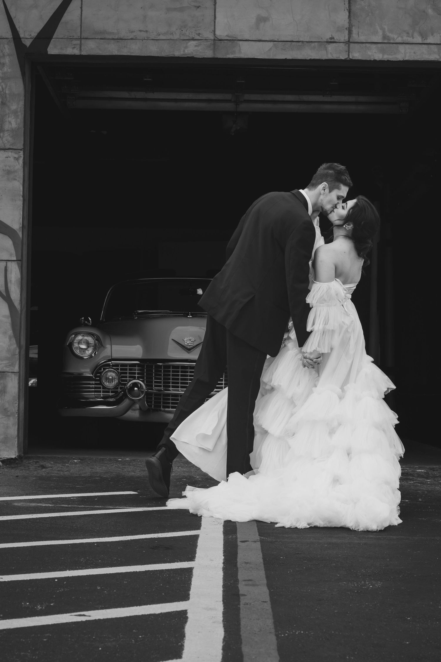Black and white photo of wedding couple kissing in front of classic car in garage.