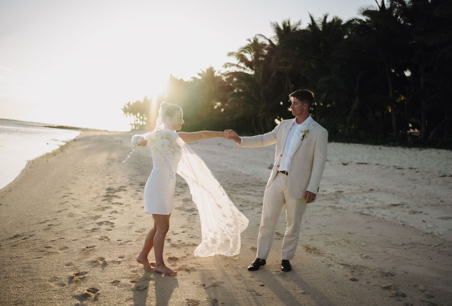 A couple in wedding attire stands on a tropical beach at sunset as a veil flows in the breeze.