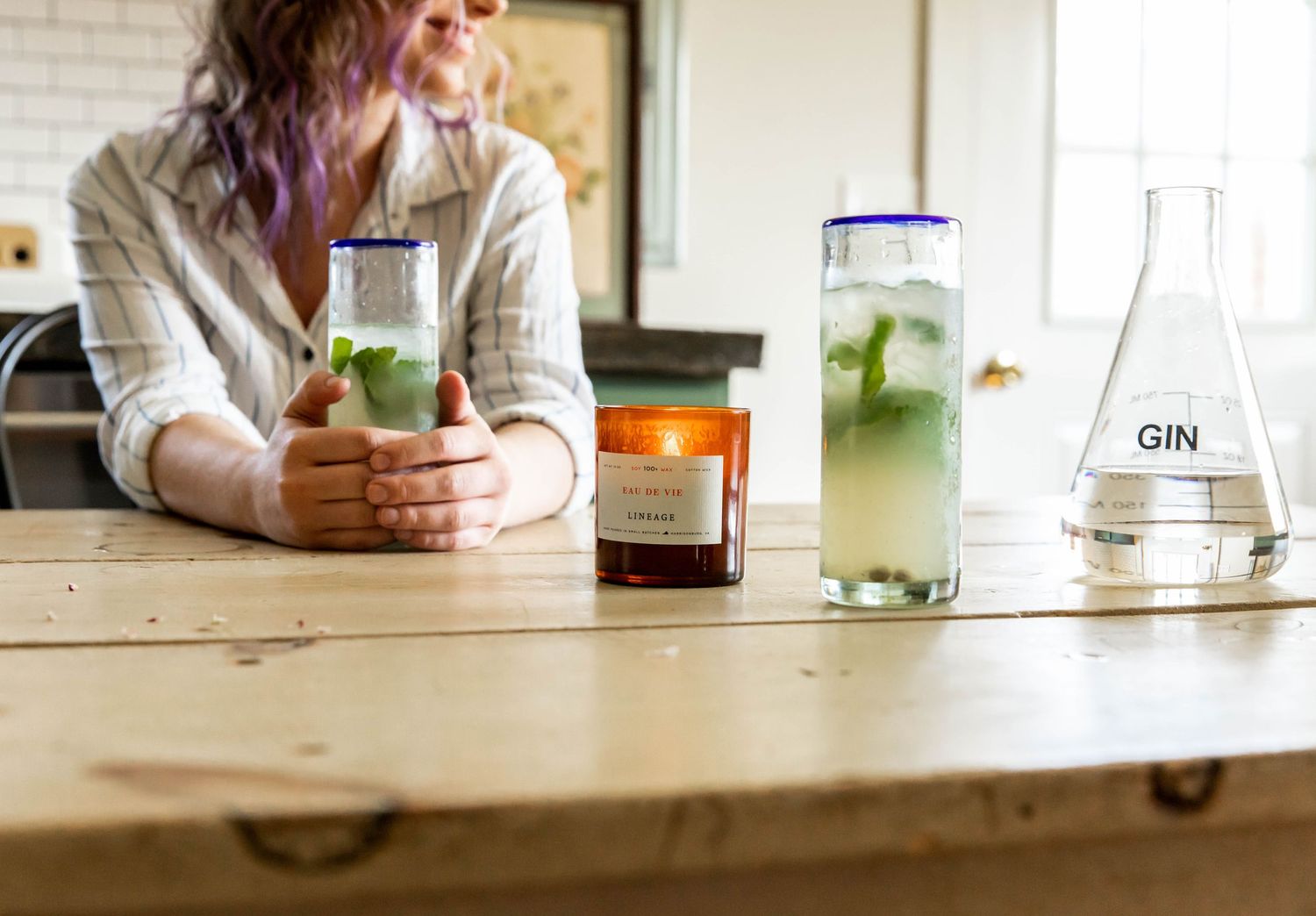 Photo of a woman sitting at a kitchen table drinking a gin and tonic, with a Lineage brand candle lit on the table