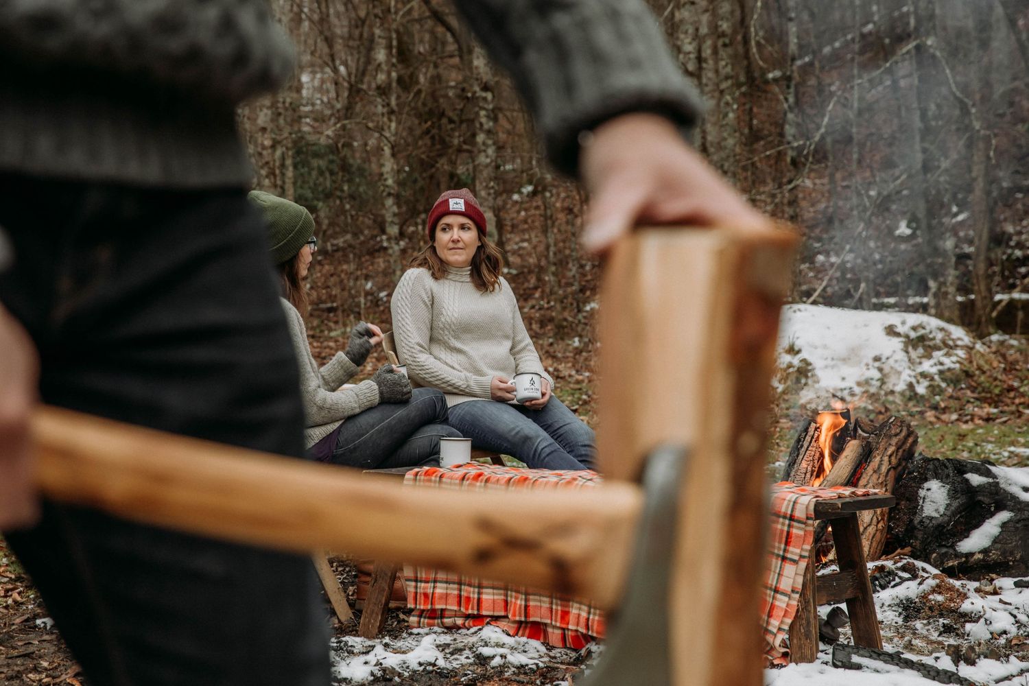 Photo of a man chopping fire wood, while women look on, drinking coffee