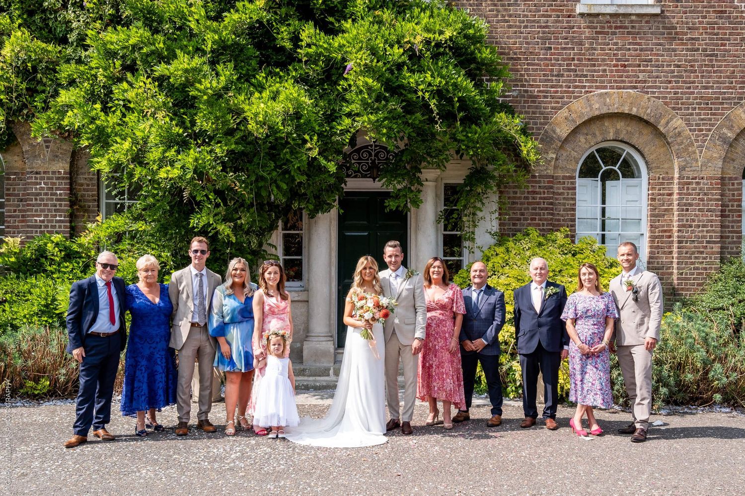 A wedding party poses for a group photo outside a historic brick building with arched windows and lush greenery.