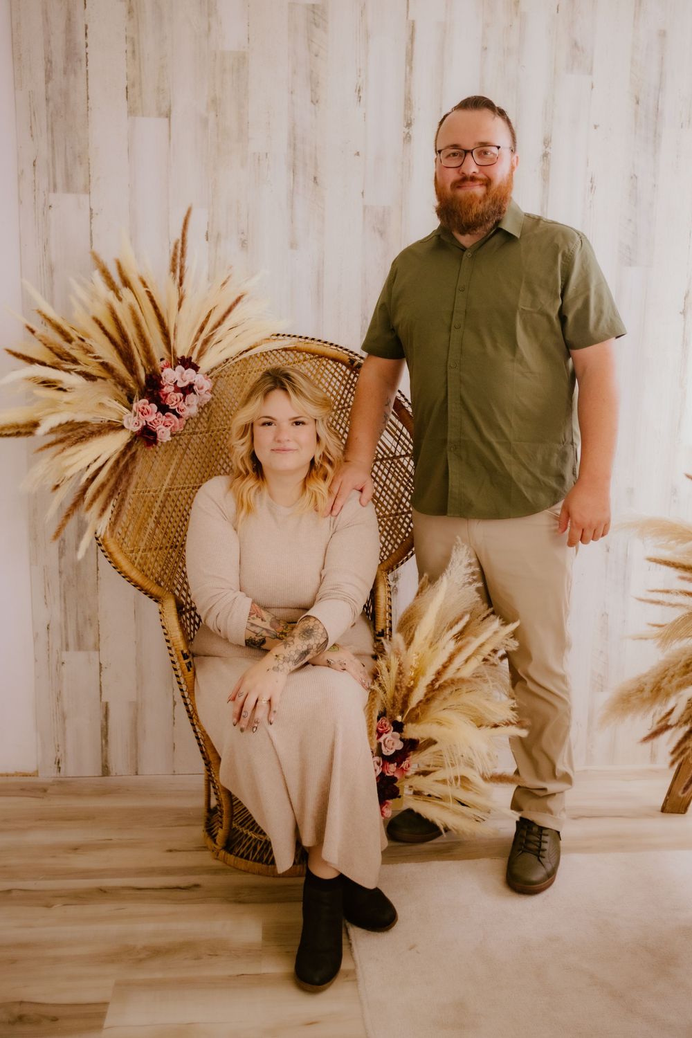 A couple poses against a wooden wall with boho decor including dried palm fronds and a wicker peacock chair.