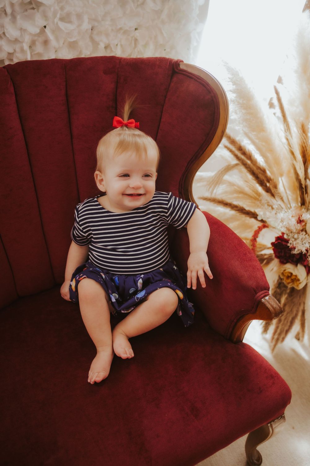 Baby in striped shirt sits on burgundy velvet couch with decorative backdrop.